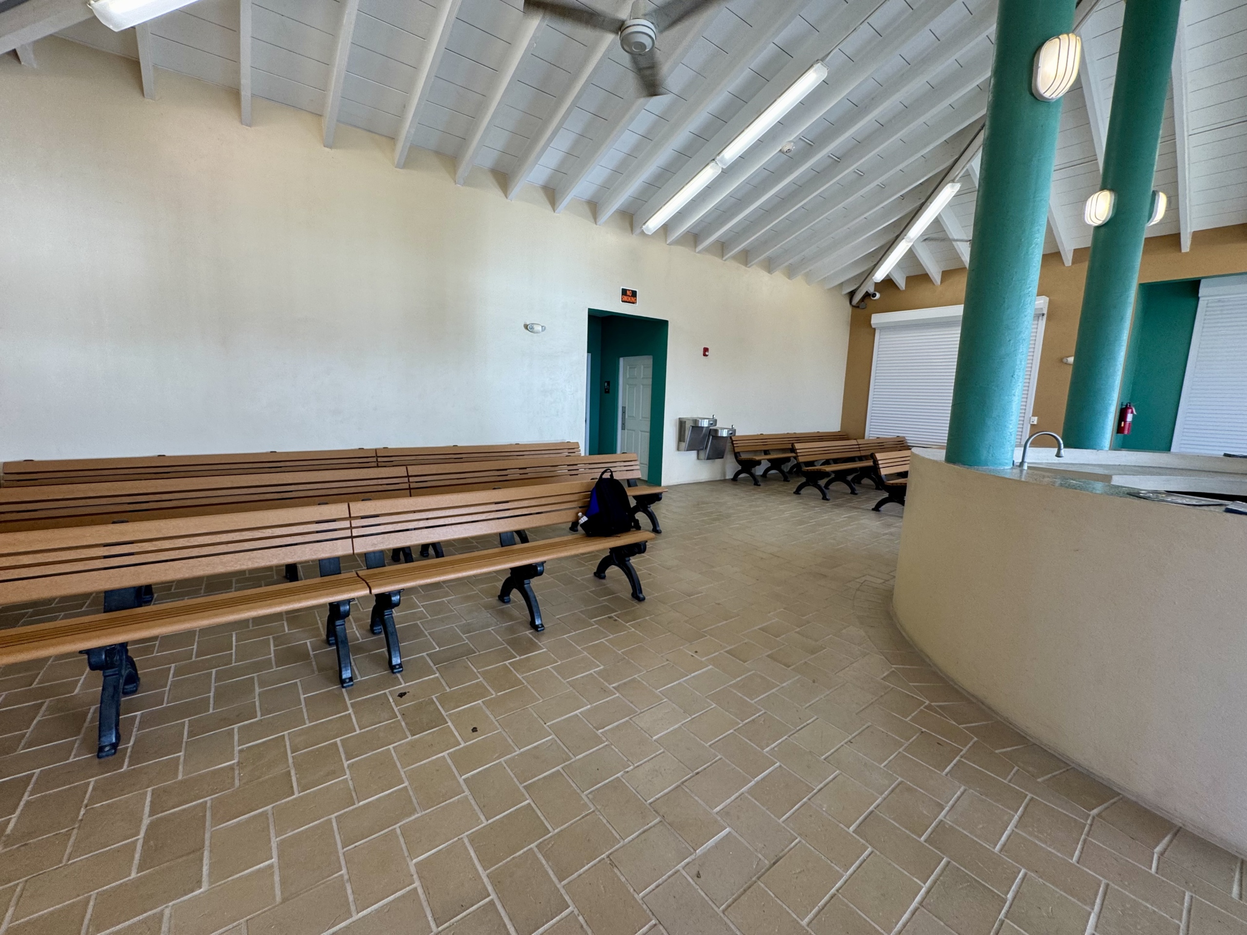 A photo of the indoor waiting area at the Gallows Bay ferry terminal in St. Croix. The space is clean and open, featuring long wooden benches with black metal legs arranged in rows. The floor is covered in tan-colored tile arranged in a herringbone pattern. The ceiling is vaulted with exposed white rafters and fluorescent lighting. Several teal-colored columns support the roof, and a small counter or service window is visible on the right. A drinking fountain and restrooms are located in the back corner. The area is well-lit and minimally decorated, offering a basic but functional waiting space for ferry passengers.