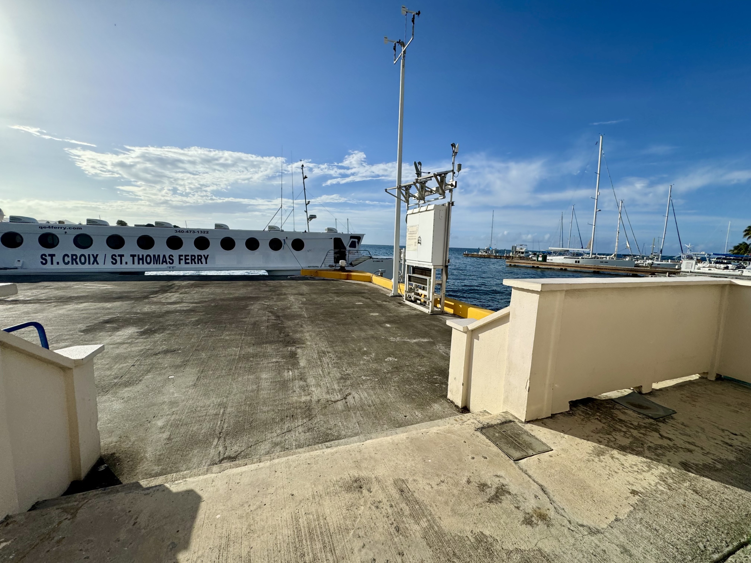 A sunny view of the QE IV Ferry docked at the terminal in Gallows Bay, St. Croix. The ferry is a white high-speed catamaran with circular windows and bold text reading “ST. CROIX / ST. THOMAS FERRY” on its side. The dock is made of smooth concrete and bordered by short beige walls. In the background, calm blue water stretches to the horizon with sailboats and yachts anchored nearby. A clear sky and bright sunlight create a vibrant, tropical setting.