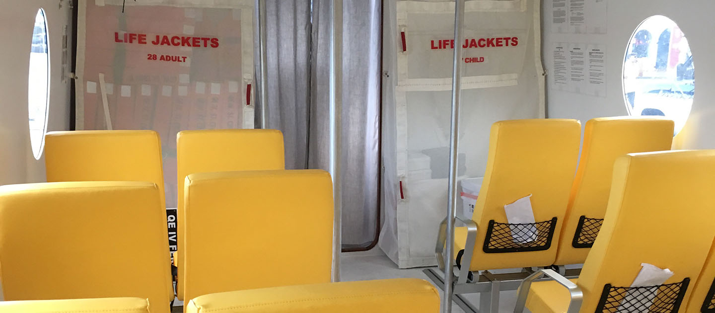 The interior of the QE IV Ferry, showing a clean and organized seating area with bright yellow cushioned chairs arranged in rows. Each seat has a small mesh pocket on the back, containing informational pamphlets or safety cards. Oval windows line the white walls, allowing natural light to brighten the space. Toward the back, labeled compartments store life jackets—“28 ADULT” on the left and “CHILD” on the right. The environment appears safe, modern, and comfortably air-conditioned for passengers.