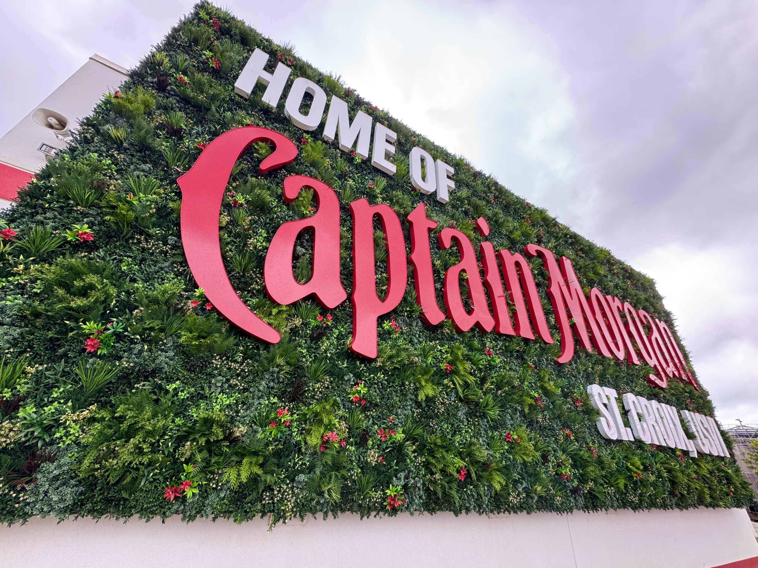 A large vertical wall covered in lush green artificial plants with small red flowers scattered throughout, creating a vibrant backdrop. Prominently displayed across the greenery is a bold, three-dimensional sign in white and red lettering. The text reads “HOME OF Captain Morgan” in large, stylized red font, with “ST. CROIX, USVI” in smaller white letters beneath it. The scene appears to be outdoors with a cloudy sky overhead, indicating overcast weather. The display is part of the Captain Morgan distillery in St. Croix, U.S. Virgin Islands.