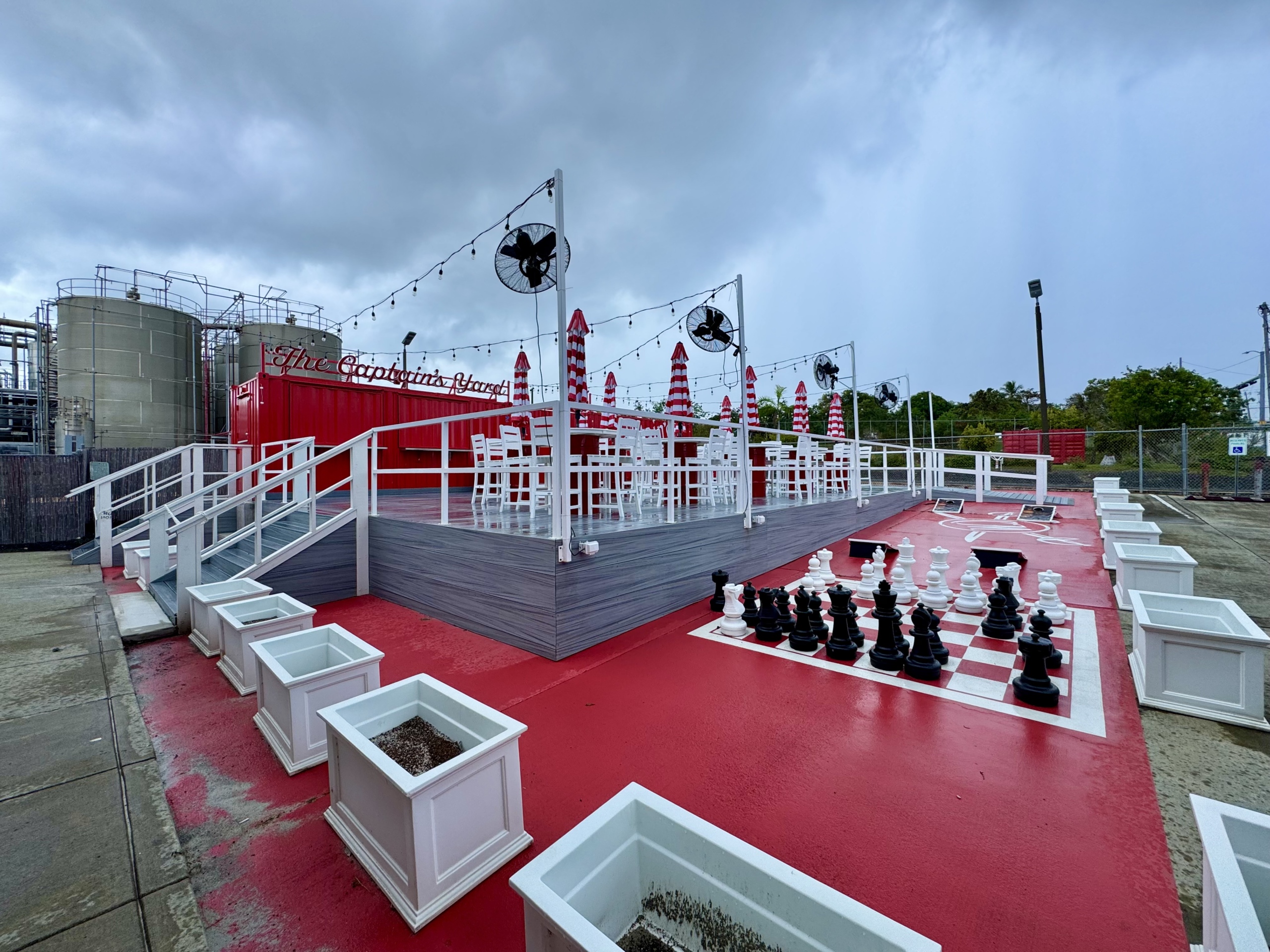An outdoor space at the Captain Morgan Distillery in St. Croix, known as “The Captain’s Yard.” The area is set up with a raised deck painted in red and gray tones, surrounded by white railings and decorated with string lights and mounted fans. Red and white striped umbrellas provide shade for high-top tables and chairs, creating a casual and festive atmosphere.
To the right of the deck, there’s a large outdoor chess board with oversized black and white chess pieces on a red-and-white painted surface. Surrounding the space are white planter boxes, most of them currently unplanted. In the background, you can see part of the distillery’s industrial operations, including large silver storage tanks and piping.
The sky is overcast, suggesting it may have recently rained or is about to rain, but the area still feels inviting and playful—a fun spot for visitors during or after a rum tasting.