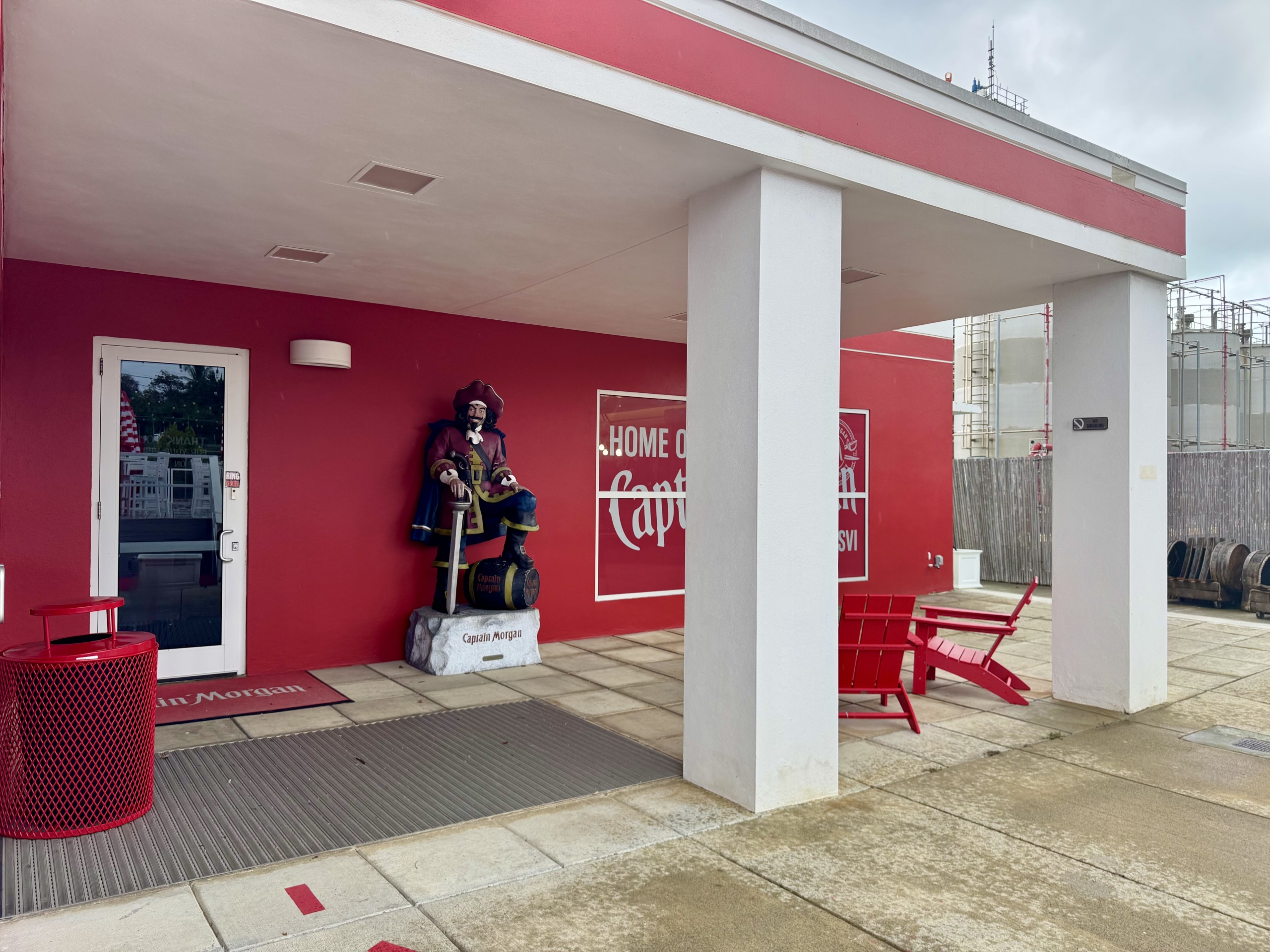 Entrance area of the Captain Morgan Visitor Center at the distillery in St. Croix, U.S. Virgin Islands. The building exterior is painted bright red with white trim and features a life-sized statue of the iconic Captain Morgan pirate, posed confidently with one foot on a barrel.
Above and behind the statue is a large wall graphic that reads “HOME OF Captain Morgan ST. CROIX, USVI”, reinforcing the brand’s connection to the island. To the right of the statue is a shaded outdoor seating area with bold red Adirondack-style chairs and a table under a white overhang supported by square columns.
Near the entrance door, there’s a red trash can and branded welcome mats. In the background, parts of the distillery operation—including tanks and equipment—are visible, blending the fun tourist vibe with the working rum production facility. The ground is wet, indicating recent rain.