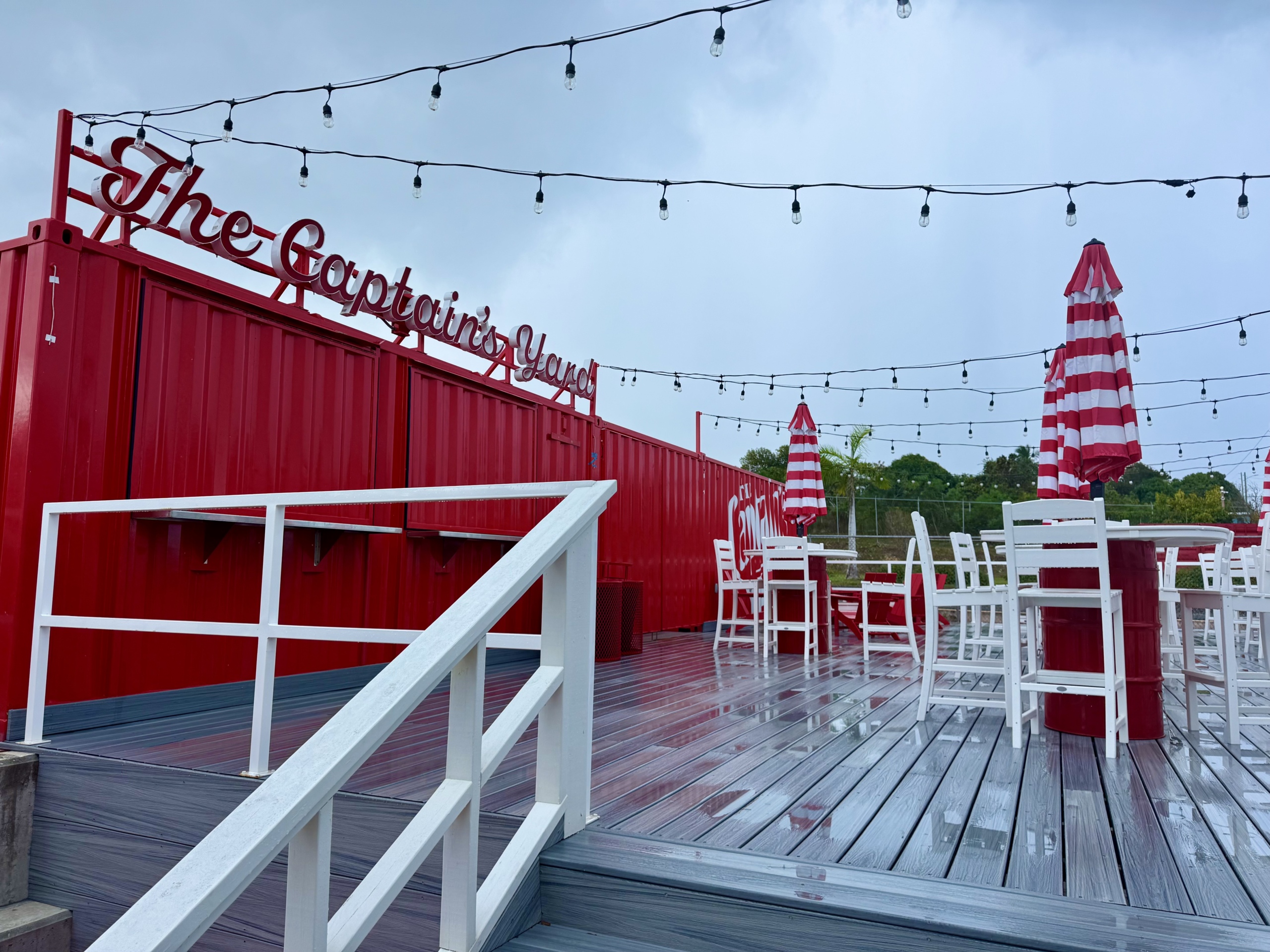 A close-up view of The Captain’s Yard, an outdoor hospitality area at the Captain Morgan Distillery in St. Croix. The space features a raised deck with string lights overhead, creating a festive and welcoming atmosphere. The deck is built with gray composite wood and enclosed by a white railing.
In the center and to the right, there are red-and-white striped umbrellas paired with white wooden high-top tables and chairs, giving it a nautical and branded theme. The back wall of the area is made of bold red shipping containers with the sign “The Captain’s Yard” in stylized script mounted at the top.
The deck appears wet from recent rain, and the sky remains overcast, adding a cool, moody contrast to the otherwise vibrant color palette. Palm trees and greenery are visible in the background, reinforcing the tropical island setting.