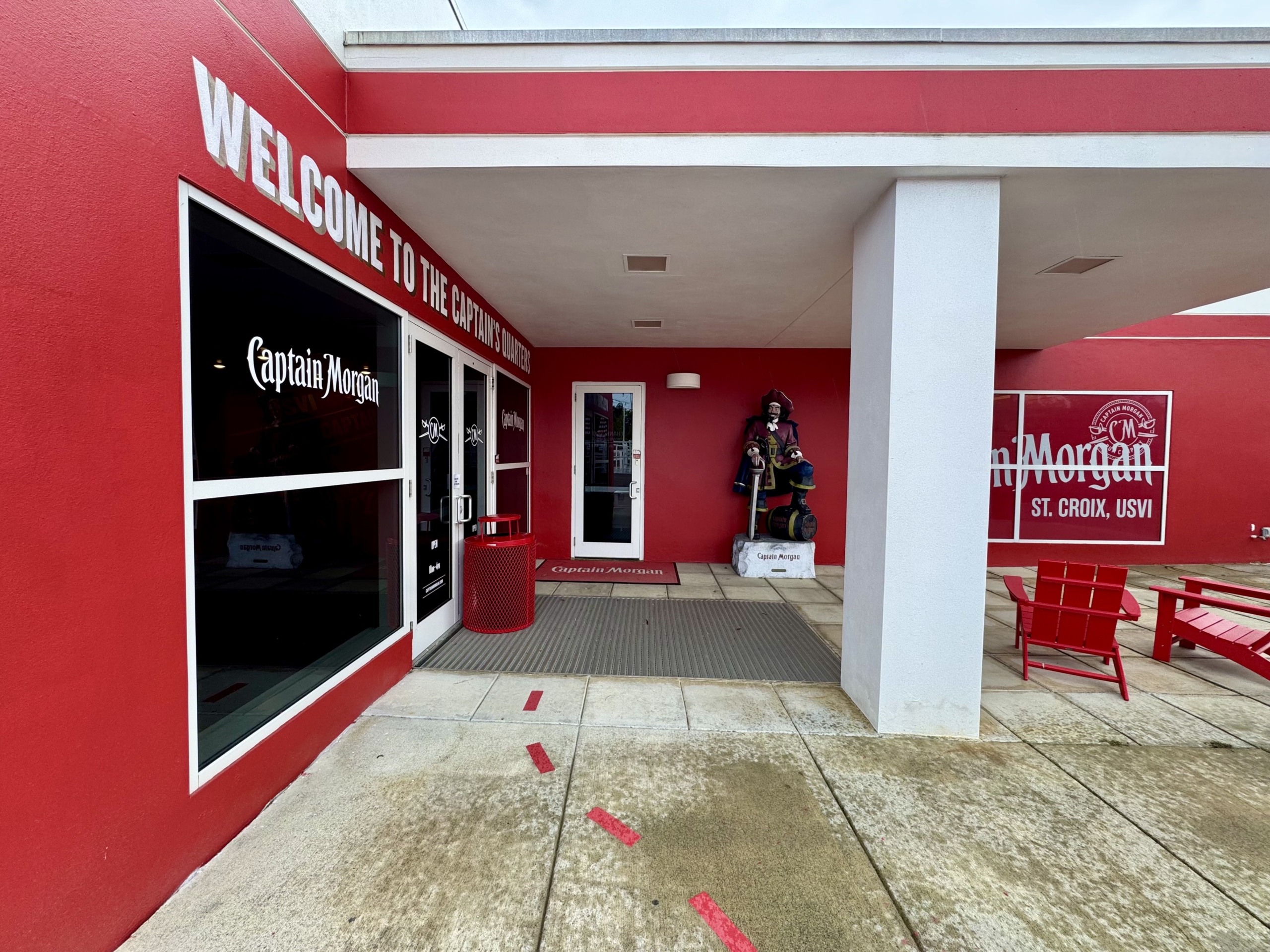 Welcoming entrance to the Captain Morgan Visitor Center at the distillery in St. Croix, U.S. Virgin Islands. The building exterior is boldly painted in red and white, with the phrase “WELCOME TO THE CAPTAIN’S QUARTERS” displayed prominently in large white letters above the windows and doors.
To the left, black-tinted windows display the Captain Morgan logo in white. Double glass doors sit at the center, flanked by a red trash can and red welcome mats branded with the Captain Morgan logo.
To the right of the entrance is a life-size statue of Captain Morgan—the brand’s signature pirate figure—standing with one foot on a rum barrel. Behind him, the building wall is decorated with the bold text “Captain Morgan ST. CROIX, USVI” in white on red. Nearby, two red Adirondack chairs and a table offer a spot to sit outside under the covered walkway. The ground is wet, indicating recent rainfall, and red directional arrows are painted on the concrete to guide visitors.