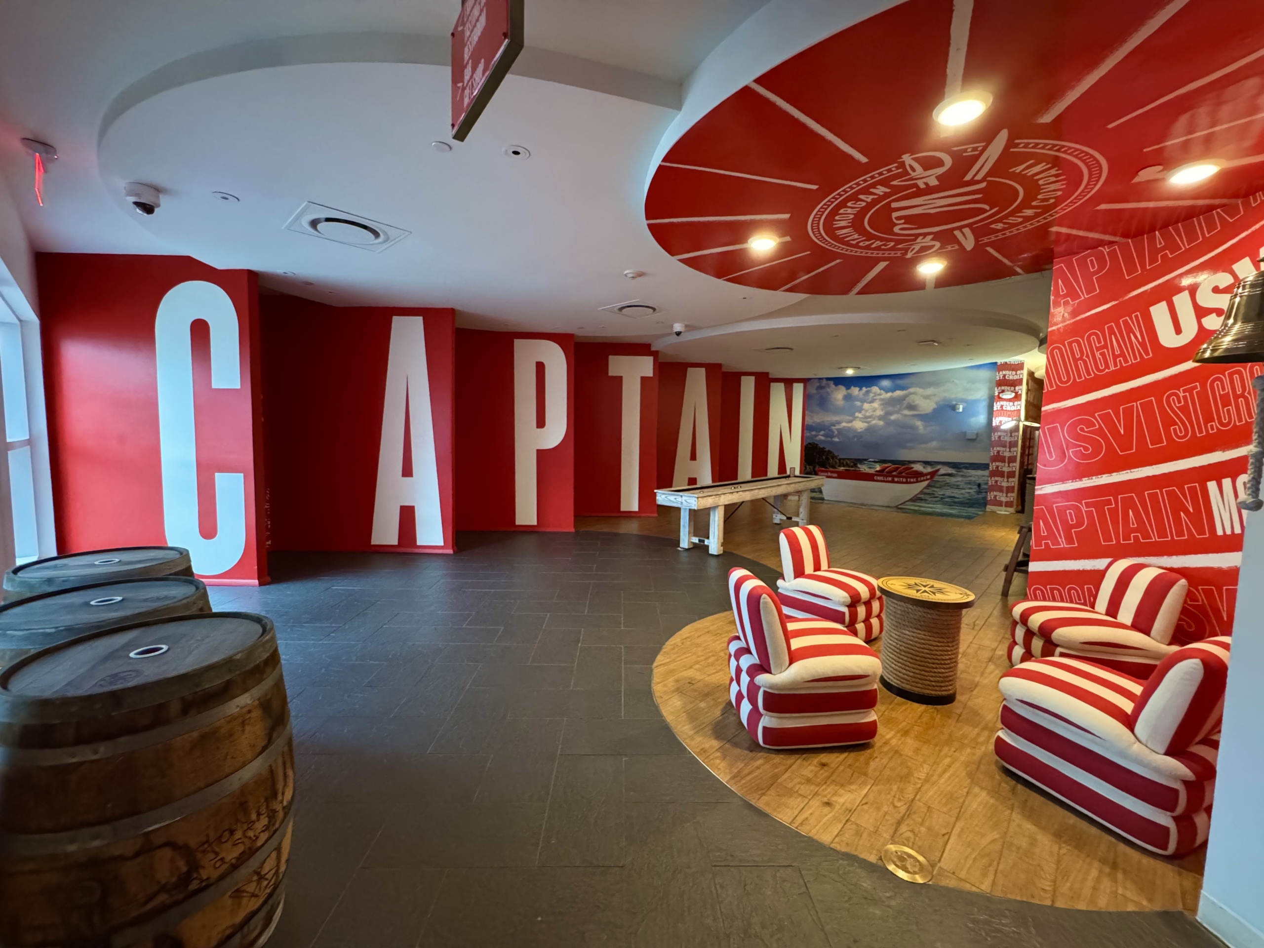 The bold and colorful interior welcome area of the Captain Morgan Visitor Center in St. Croix, U.S. Virgin Islands. The space features a striking red-and-white color scheme and is designed to be fun, energetic, and highly branded.
On the far wall, oversized white letters on a red background spell out the word “CAPTAIN” in block format, creating a perfect photo-op backdrop. To the right, a cozy lounge area includes four plush, red-and-white striped chairs arranged around a small round table wrapped in nautical rope, sitting atop a circular wood-toned platform.
The ceiling above this area is painted red and features the Captain Morgan emblem, illuminated by recessed lights. Along another wall, repeated text reads “CAPTAIN MORGAN USVI ST. CROIX” in alternating directions, adding to the branded atmosphere. There is also a large scenic photo mural of a red Captain Morgan boat on the water, further reinforcing the tropical, adventurous feel. In the foreground to the left, a row of aged wooden rum barrels nods to the distillery’s craft and heritage.