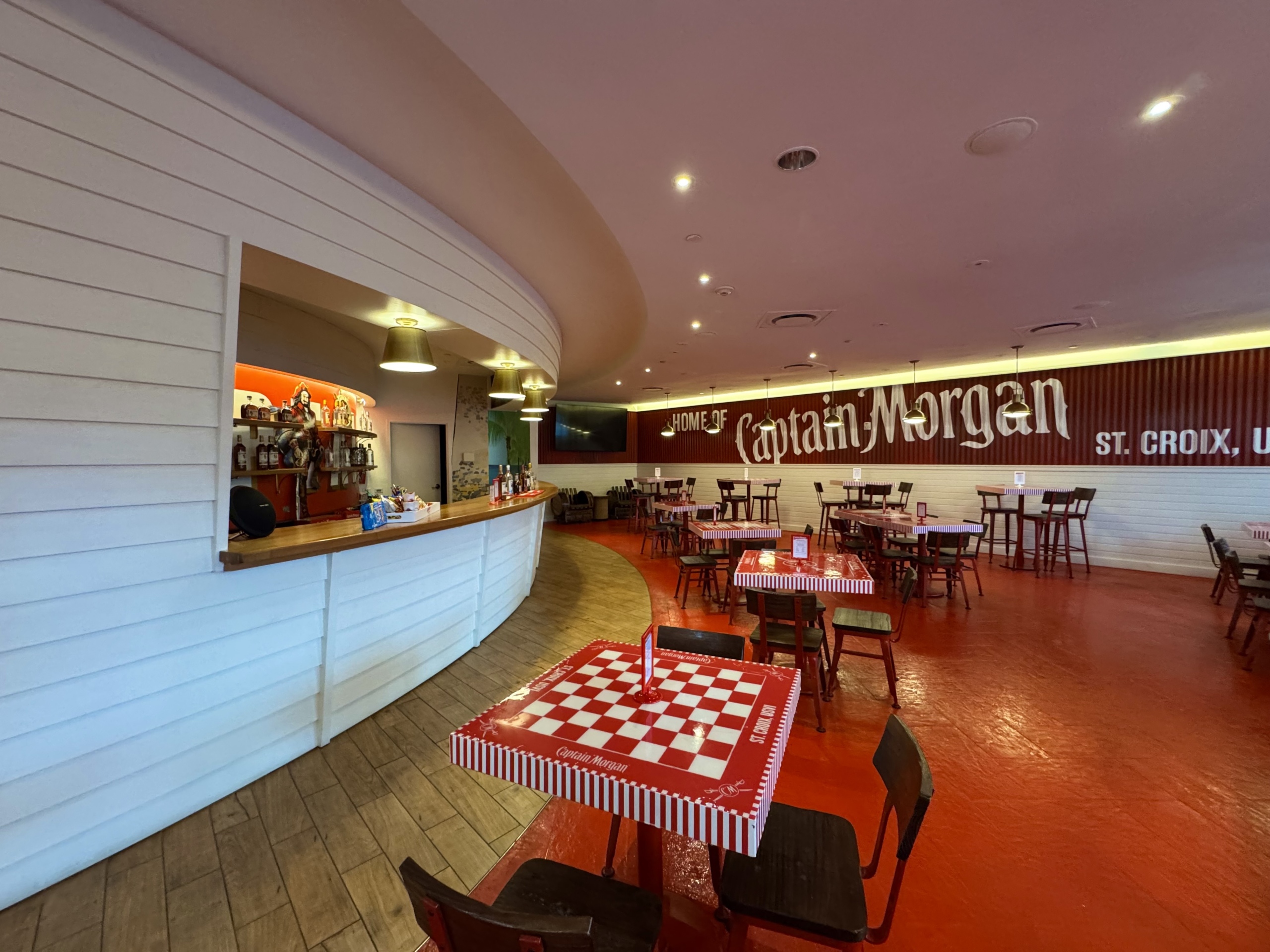 Lively indoor tasting area at the Captain Morgan Distillery in St. Croix, U.S. Virgin Islands. The room is spacious and warmly lit, combining elements of a bar and a casual dining or lounge setting.
On the left side of the image is a white-paneled curved bar with a wooden countertop. Behind the bar, a brightly lit shelf displays a variety of Captain Morgan rum bottles and branded memorabilia, including a pirate figurine.
The floor is painted a rich red, matching the color of the square checkerboard-patterned tables which are striped and branded with the Captain Morgan logo. Wooden chairs are arranged around each table, offering plenty of seating for groups and individuals.
On the far wall, bold white text reads “HOME OF Captain Morgan ST. CROIX, USVI”, displayed over red corrugated metal and white shiplap siding. The overall ambiance feels welcoming, fun, and distinctly themed for a rum tasting experience.
