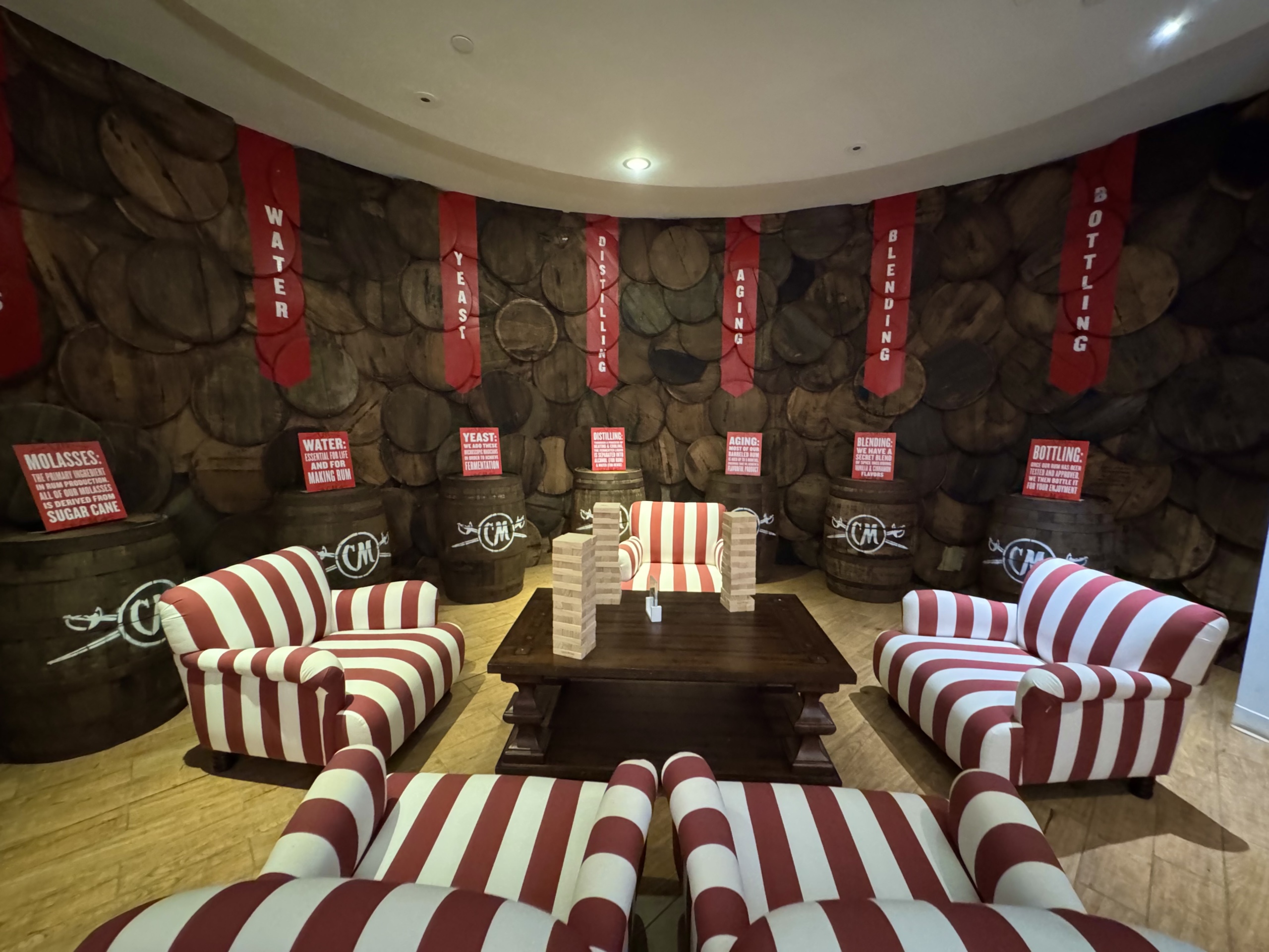 A cozy and educational lounge area inside the Captain Morgan Visitor Center in St. Croix, U.S. Virgin Islands. The backdrop is a mural of stacked wooden barrels, each symbolizing a part of the rum-making process. Vertical red banners with white text hang across the mural, highlighting each stage: Water, Yeast, Distilling, Aging, Blending, Bottling.
In front of the mural are several real wooden barrels displaying educational signs with brief explanations of each step in the rum production process. Examples include: “Molasses: The primary ingredient used in making rum, as a byproduct from sugar cane.” “Aging: Rum is stored in oak barrels to enhance flavor.”
In the center of the space, a large wooden coffee table is surrounded by six plush red-and-white striped chairs, creating a nautical, branded look. A game of giant Jenga is set up on the table, inviting visitors to relax and interact while learning. The area is warmly lit and laid out on wood-toned flooring, blending comfort with storytelling in a fun and engaging setting.