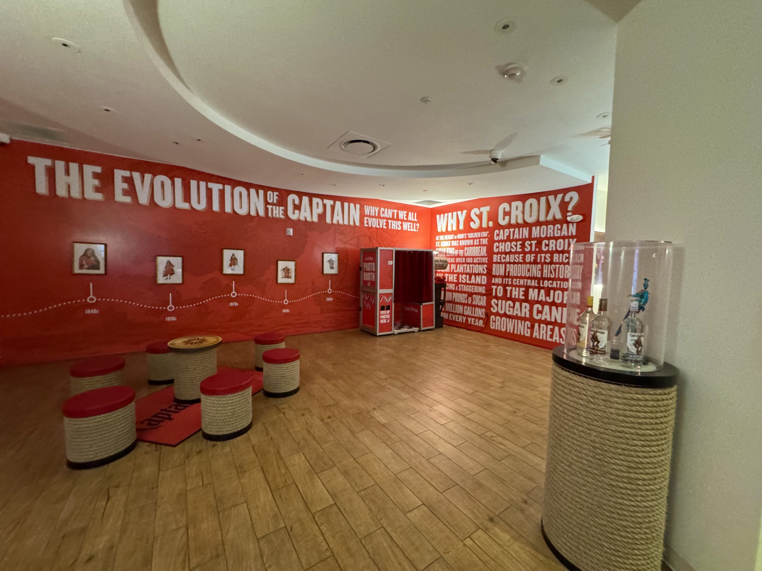 A vibrant exhibit space inside the Captain Morgan Visitor Center in St. Croix, USVI, focused on the brand’s heritage and local connection.
On the left wall, bold white text reads “THE EVOLUTION OF THE CAPTAIN,” displaying a visual timeline of the Captain Morgan mascot’s changing appearance from the 1940s to modern day. Framed illustrations of the character are spaced out along a dotted path labeled by decade, showing how the branding and costume evolved over time.
To the right, the adjacent wall features the large heading “WHY ST. CROIX?” in white block letters. The accompanying text explains that Captain Morgan chose St. Croix because of the island’s rich rum-producing history, fertile land for sugar cane, and central location in the Caribbean. The paragraph is visually striking and uses red and white for high contrast.
In the center of the room, a round seating area includes stools wrapped in nautical rope with red cushions, and a small round table bearing a compass rose design. To the far right, a cylindrical display case holds a trio of Captain Morgan bottles, highlighting product variety. Just behind the exhibit is a small photo booth, offering a fun souvenir opportunity for visitors.
The overall setting is polished, educational, and interactive—celebrating both the legacy of Captain Morgan and its island roots.