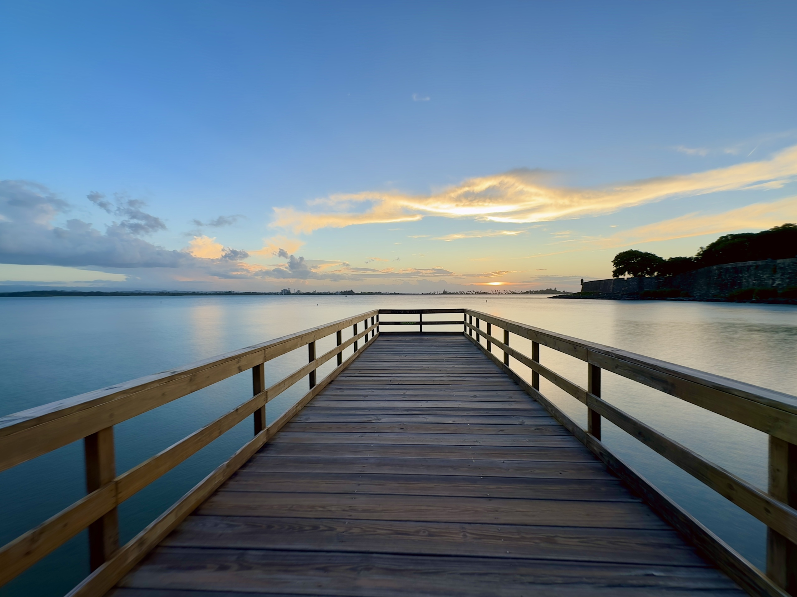 A tranquil sunset scene viewed from a wooden pier extending out over calm, reflective water. The pier’s warm-toned railings lead the eye toward the distant horizon, where soft golden light glows beneath streaks of pastel clouds. On the right side of the frame, the silhouette of a stone fort and trees gently curves along the shoreline, part of Old San Juan, Puerto Rico. The sky transitions from blue to warm orange and pink as the sun sets, casting a peaceful, serene atmosphere across the scene.
