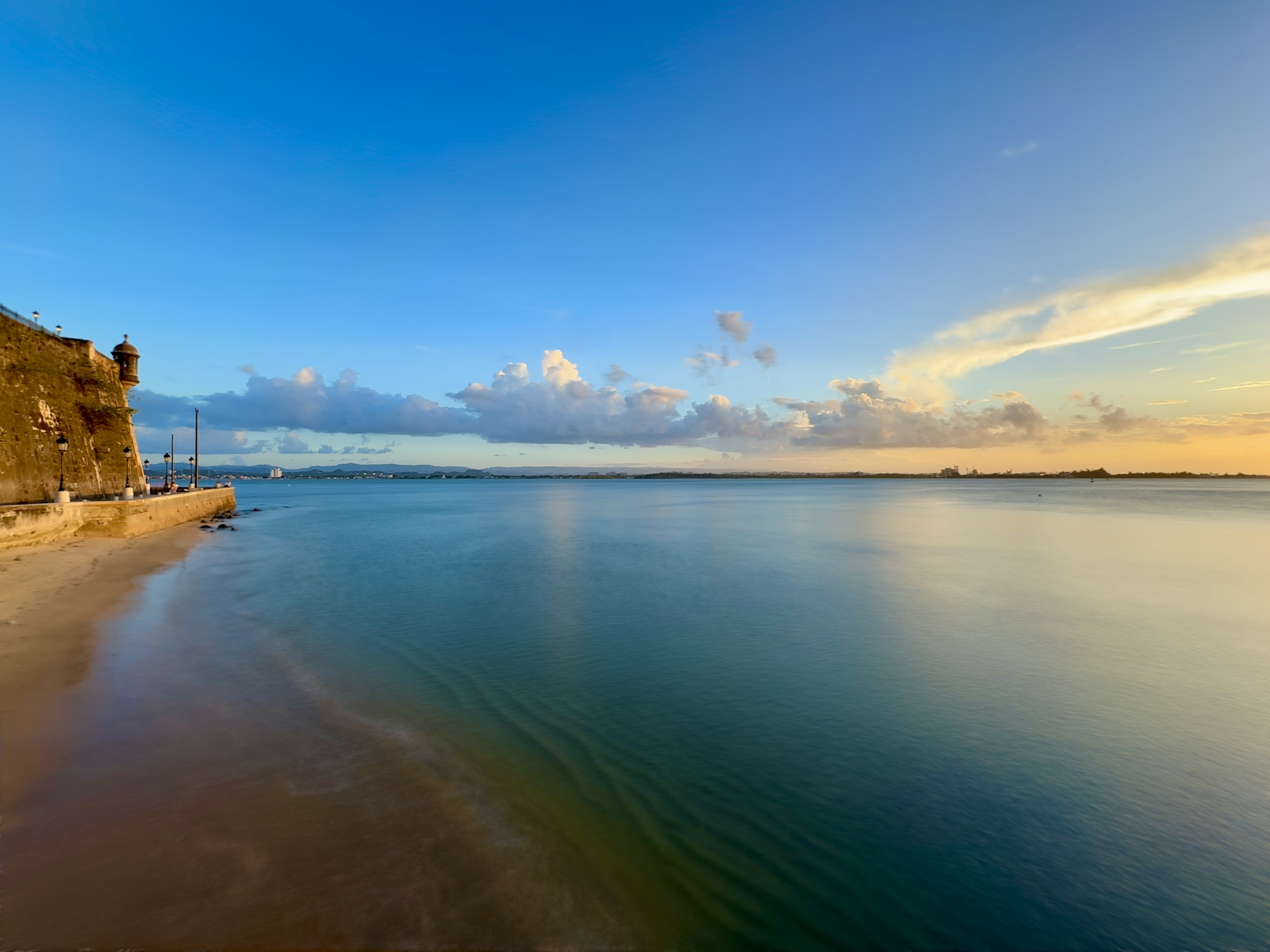 A peaceful coastal view at sunset, with calm, glassy water stretching across the frame. On the left, a historic stone fort wall—part of Old San Juan’s iconic city walls—extends along the shoreline, with a small guard tower (garita) perched at the corner. Lampposts line the pathway beneath the fort, casting soft light. The sandy shore curves gently along the base of the wall. The sky is a blend of soft blue and warm golden hues, with scattered clouds catching the glow of the setting sun on the right. The atmosphere is calm, historic, and reflective.
