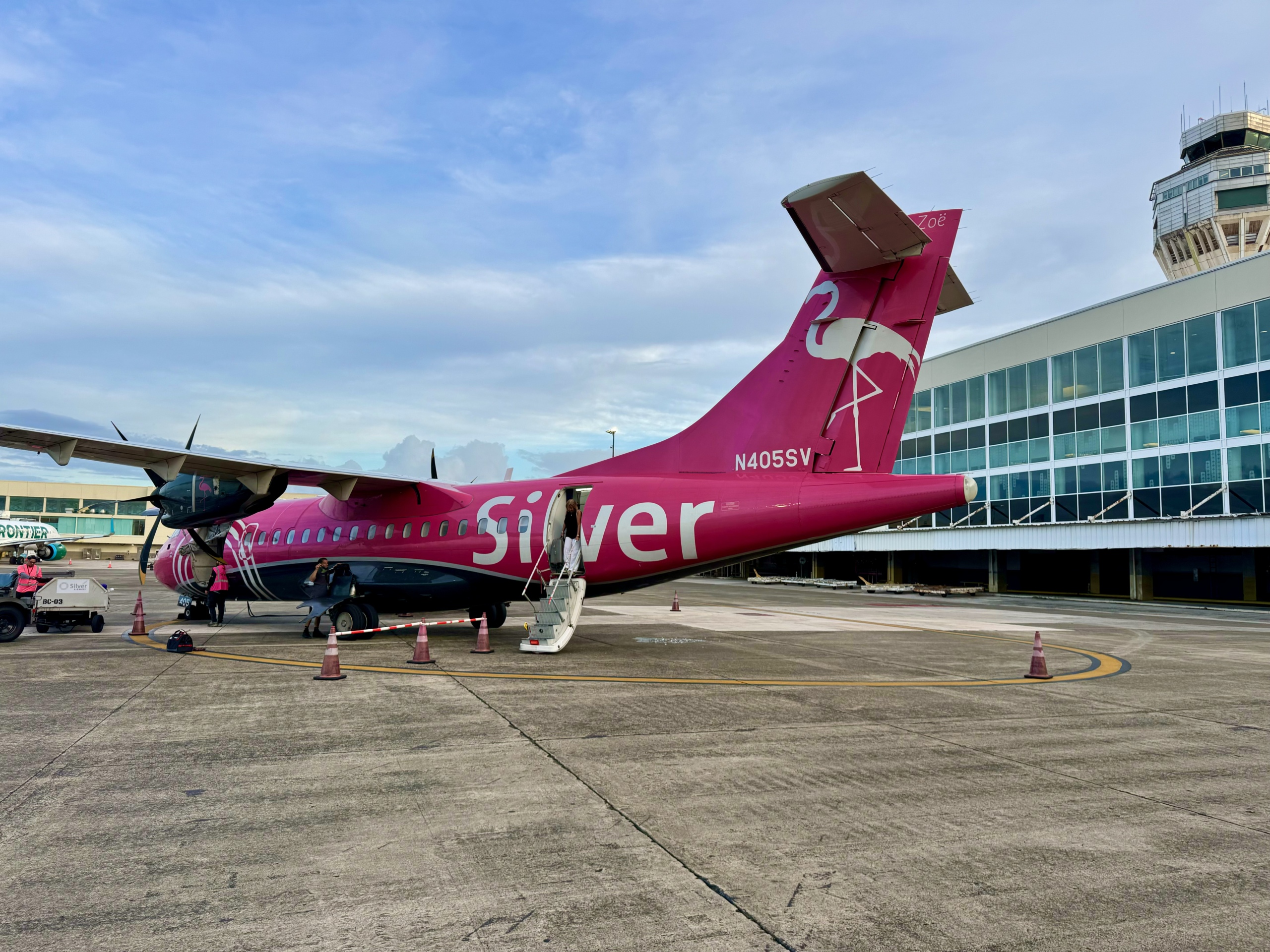 A bright magenta and white Silver Airways aircraft is parked on the tarmac at an airport terminal. The turboprop plane features the name “Silver” in large white letters along the fuselage and a stylized white flamingo graphic on the tail, with the name “Zoë” written near the top. The aircraft’s registration number, N405SV, is visible near the tail. Several workers in pink uniforms are servicing the aircraft, with a stairway extended for boarding. The terminal building in the background has large glass windows and a control tower rises above it. The scene is calm with soft lighting, suggesting early morning or late afternoon.