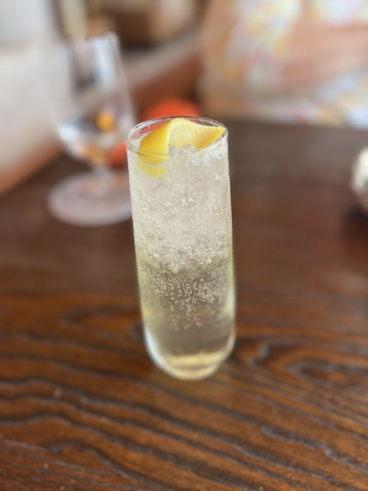 A close-up photo of a tall, clear glass filled with a sparkling, pale-yellow drink garnished with a lemon twist. The drink sits on a dark wooden table with a subtle grain pattern. In the blurred background, there is a partially visible water glass and the colorful outline of a person wearing a pastel-patterned outfit. The image evokes a refreshing and upscale dining experience.