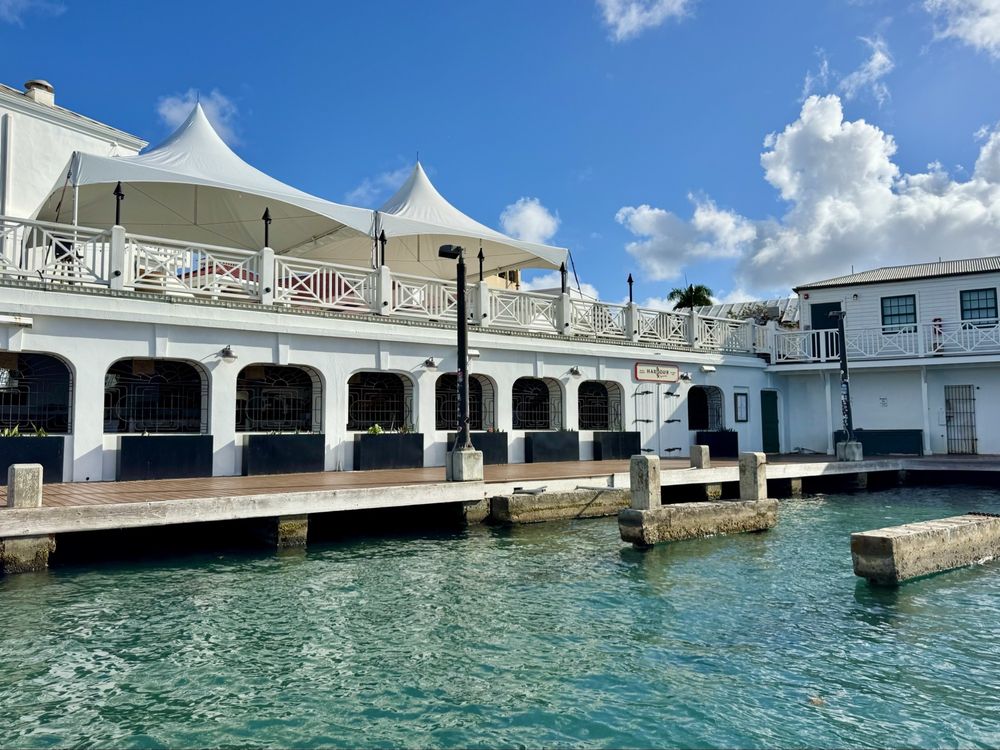 A scenic view of Harbour Prime restaurant in Christiansted, St. Croix, taken from across the turquoise waters of the harbor. The white, two-story building features arched windows and a wooden boardwalk at the water’s edge. On the upper level, white tents provide shade for the outdoor dining area, framed by white railings. The sky above is bright blue with scattered clouds, giving the scene a clean, coastal atmosphere.
