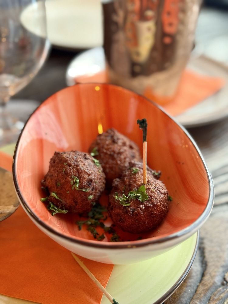 A close-up image of a dish containing three golden-brown fritters garnished with chopped herbs, served in an orange, oval-shaped bowl. One fritter is pierced with a cocktail pick. The bowl rests on a light green plate with an orange napkin underneath. In the softly blurred background, a water glass and a decorative tumbler sit on a wooden table, adding to the upscale, casual dining ambiance.