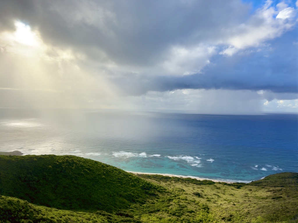 Panoramic view overlooking Jack and Isaac Bay on St. Croix, with emerald-green hills, turquoise shoreline, and dramatic sunbeams breaking through tropical storm clouds.