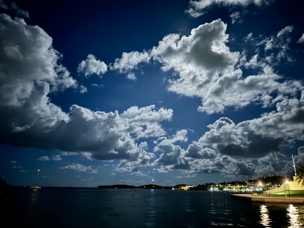 Night view from the Christiansted Boardwalk overlooking Christiansted Harbor under a dramatic moonlit sky, with illuminated clouds, sailboats at anchor, calm Caribbean waters, and the waterfront lights of St. Croix reflected on the sea.