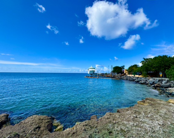 View of Frederiksted's waterfront in St. Croix with crystal-clear Caribbean water, a cruise ship docked at the Frederiksted Pier, and a bright blue tropical sky seen from the rocky shoreline.