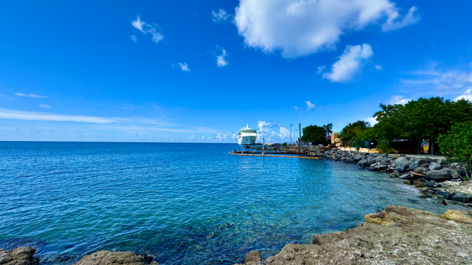 View of Frederiksted's waterfront in St. Croix with crystal-clear Caribbean water, a cruise ship docked at the Frederiksted Pier, and a bright blue tropical sky seen from the rocky shoreline.