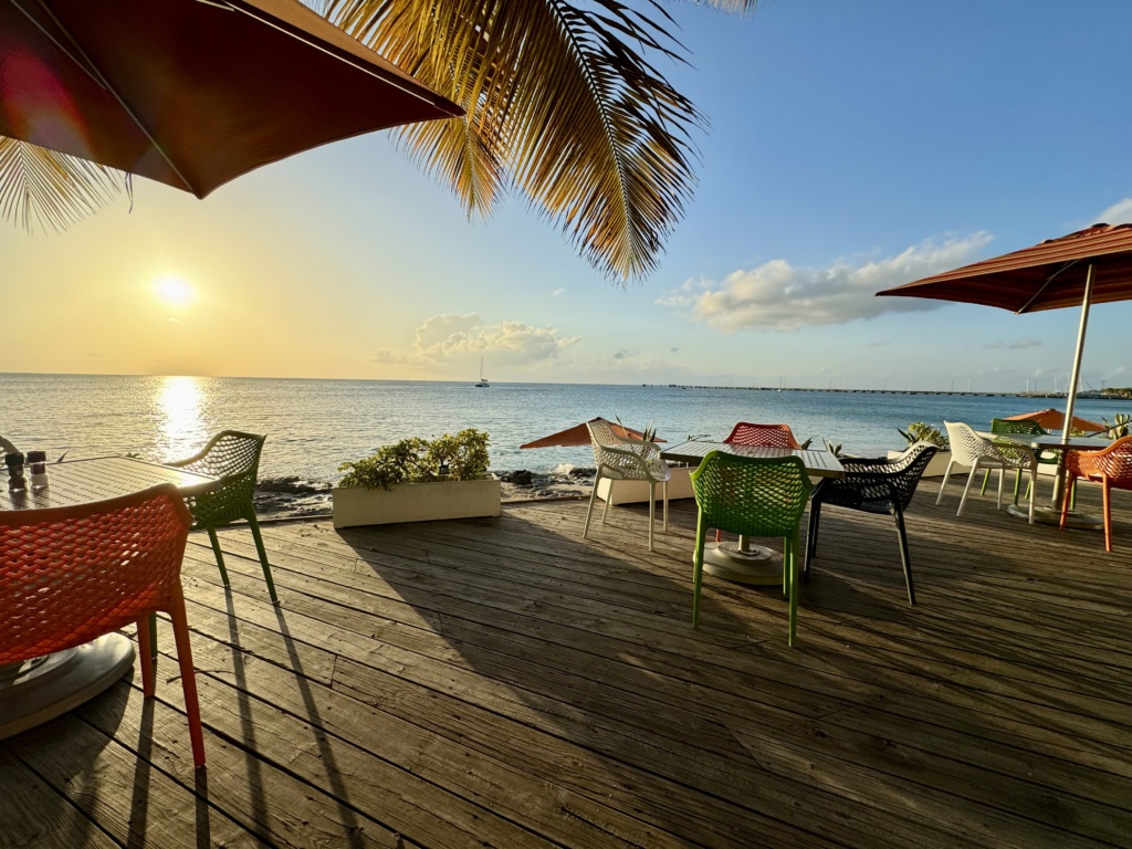 Sunset over the Caribbean Sea viewed from the outdoor deck at The Fred in Frederiksted, St. Croix, with colorful patio seating, palm fronds, and calm ocean waters glowing in the evening light.