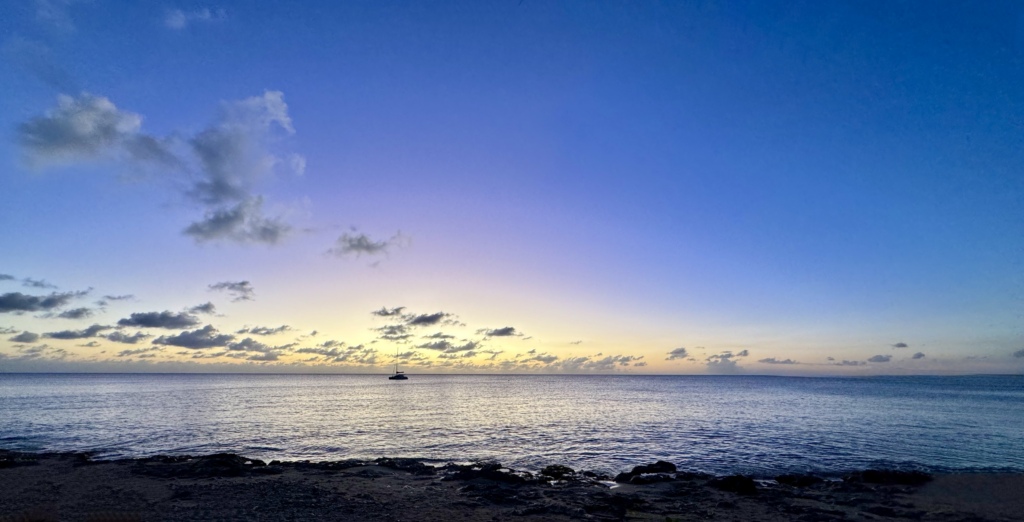 A tranquil Caribbean sunset on St. Croix with calm ocean waters, a lone sailboat anchored on the horizon, and a soft blue, purple, and golden sky reflected across the sea.