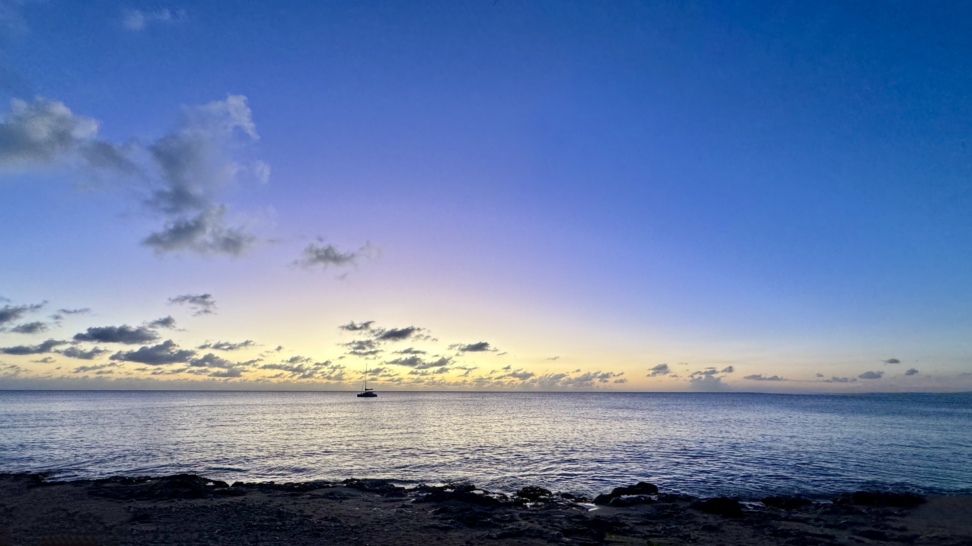 A tranquil Caribbean sunset on St. Croix with calm ocean waters, a lone sailboat anchored on the horizon, and a soft blue, purple, and golden sky reflected across the sea.